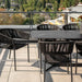 Close Up View Of The Left Side of a Black Table Next To Several Afton Outdoor Dining Chairs With Black Cord And Black Frame On A Patio