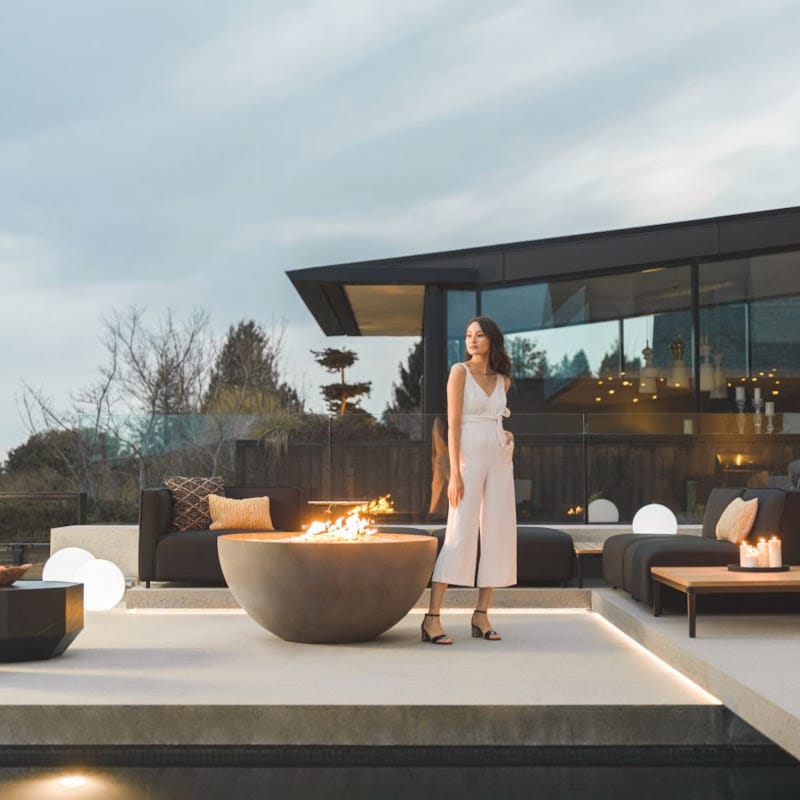 Woman Standing Next To a Fire Pit In Front Of The Arya Outdoor Modular Sectional Sofa In Acadia Color